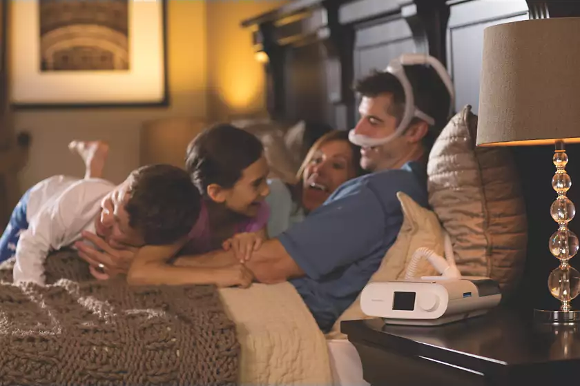 A Philips Respironics DreamStation Auto CPAP user with the machine by the bedside wearing DreamWear Nasal Mask and lying in bed with two kids and his wife