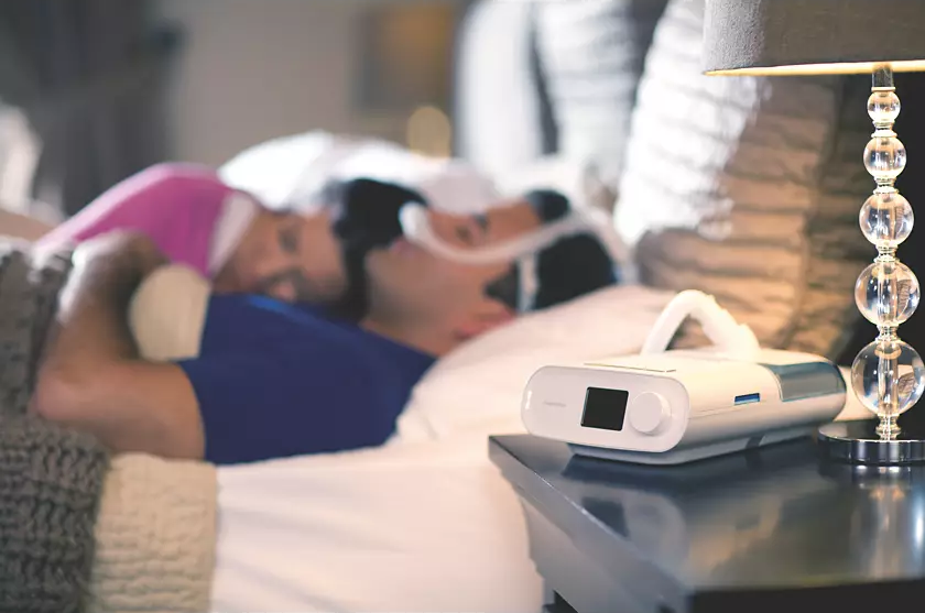 A Philips Respironics DreamStation Auto CPAP user with the machine by the bedside wearing DreamWear Nasal Mask and lying in bed with his wife